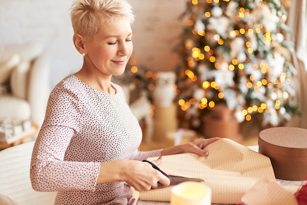 Vacanze, celebrazione e concetto di vacanze. Elegante bella donna matura con i capelli corti in posa in soggiorno decorato con albero di Natale, taglio di confezioni regalo con le forbici