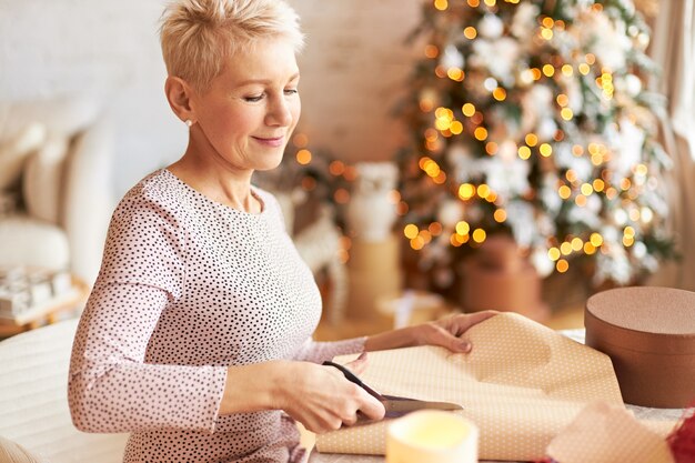 Vacanze, celebrazione e concetto di vacanze. Elegante bella donna matura con i capelli corti in posa in soggiorno decorato con albero di Natale, taglio di confezioni regalo con le forbici