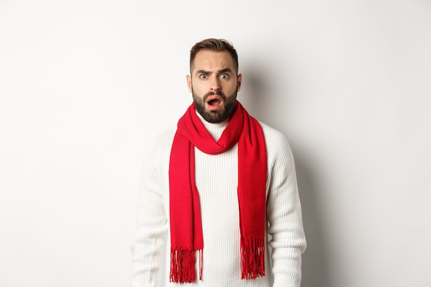 Vacanze di Natale e concetto di celebrazione. Ragazzo barbuto confuso che fissa qualcosa di strano, in piedi con sciarpa rossa e maglione, sfondo bianco.