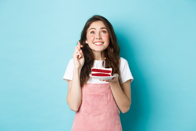 Vacanze e celebrazione. Speranzosa ragazza che festeggia il compleanno, alzando lo sguardo e incrociando le dita per buona fortuna, esprimendo desiderio sulla candela nella torta, in piedi su sfondo blu.