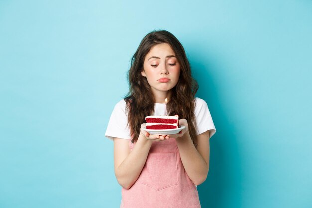 Vacanze e festeggiamenti triste ragazza carina che festeggia il suo compleanno da sola con in mano una torta sul piatto con uno...