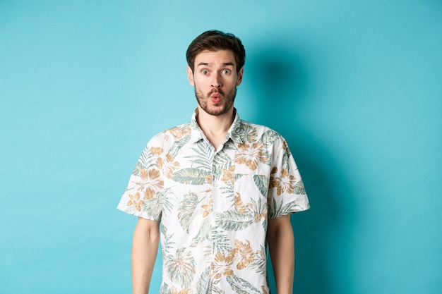 Vacanze estive. Il turista sorpreso dice wow e fissa la telecamera, guardando il fantastico promo, in piedi in camicia hawaiana su sfondo blu.