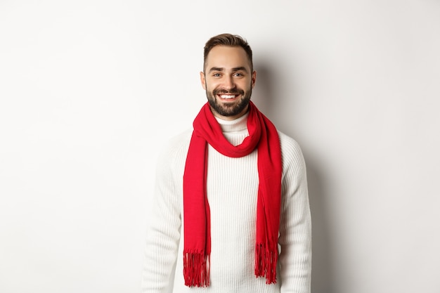 Vacanze invernali. Bell'uomo adulto con sciarpa rossa che guarda felice la telecamera, in piedi con un maglione su sfondo bianco