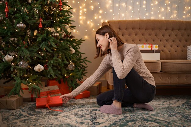 Vacanze invernali, celebrazione e concetto della gente - Chiuda in su della donna che mette presente sotto l'albero di Natale