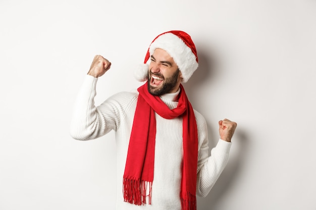 Vacanze invernali e concetto di festa di Capodanno. Bell'uomo barbuto con cappello da Babbo Natale vincendo il premio, raggiunge l'obiettivo e festeggia, facendo pompa a pugno e dicendo sì, sfondo bianco.