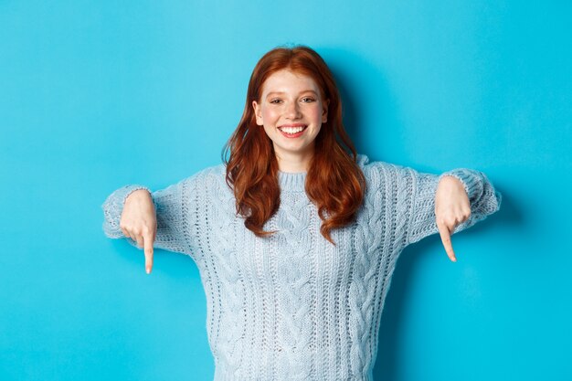 Vacanze invernali e concetto di persone. Donna rossa allegra in maglione che punta le dita verso il basso, sorride compiaciuta alla telecamera, mostrando promo, sfondo blu