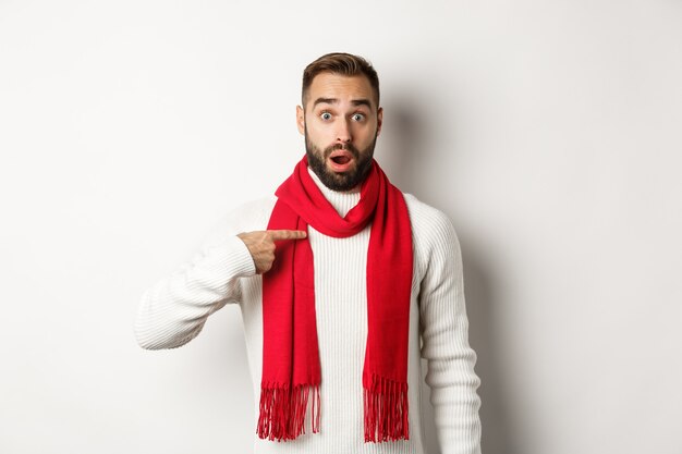 Vacanze invernali e concetto di shopping. Ragazzo sorpreso e confuso che indica se stesso, essendo scelto, in piedi con sciarpa rossa e maglione su sfondo bianco