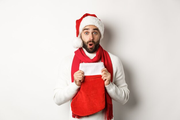 Vacanze invernali e concetto di shopping. Uomo sorpreso con cappello da Babbo Natale che riceve un regalo in un calzino di Natale, sembra stupito, in piedi su sfondo bianco