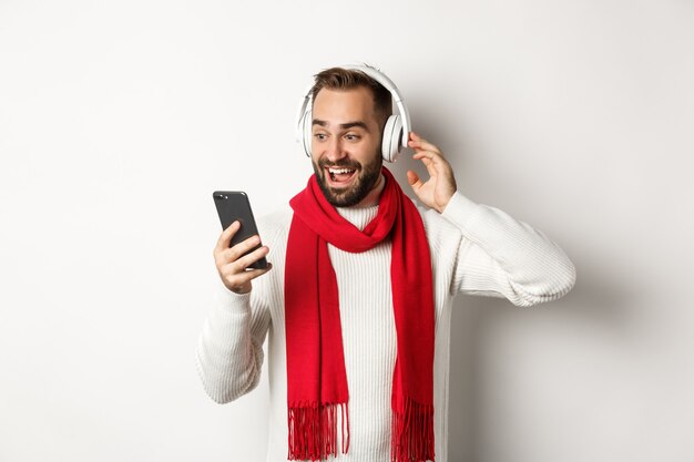 Vacanze invernali e concetto di tecnologia. Uomo felice che ascolta musica in cuffia, guardando stupito lo schermo del cellulare, in piedi su sfondo bianco.