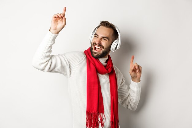 Vacanze invernali e concetto di tecnologia. Uomo felice che balla con la musica in cuffia, in piedi su sfondo bianco in maglione