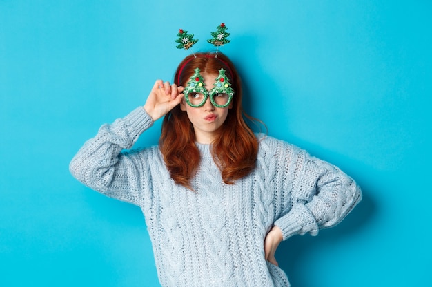 Vacanze invernali e concetto di vendite di Natale. Bellissima modella rossa che celebra il nuovo anno, indossa una fascia e occhiali divertenti, sorridente sciocco, sfondo blu.