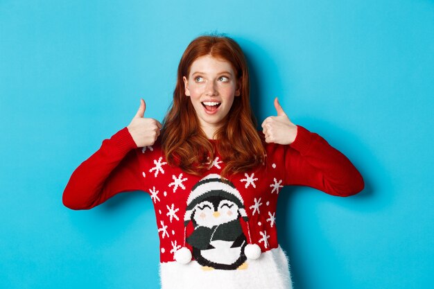 Vacanze invernali e concetto di vigilia di Natale. Bella ragazza rossa con un maglione natalizio, che celebra il nuovo anno, che mostra i pollici in su e guarda l'angolo in alto a sinistra, sfondo blu.