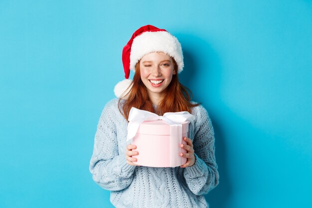 Vacanze invernali e concetto di vigilia di Natale. Carina ragazza rossa in maglione e cappello di Babbo Natale, con in mano un regalo di Capodanno e guardando la telecamera, in piedi su sfondo blu