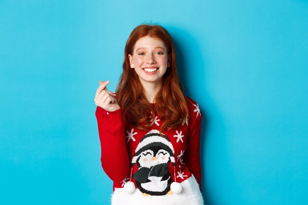 Vacanze invernali e concetto di vigilia di Natale. Carina ragazza rossa sorridente con un maglione natalizio, che mostra il segno del cuore e augura buon anno, in piedi su sfondo blu.