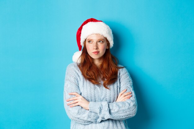 Vacanze invernali e concetto di vigilia di Natale. Donna rossa premurosa con cappello e maglione da Babbo Natale, guardando a sinistra e meditando, facendo piani natalizi, in piedi su sfondo blu.