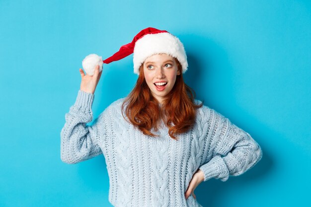 Vacanze invernali e concetto di vigilia di Natale. Sciocco ragazza rossa con le lentiggini, toccando il suo cappello da Babbo Natale e pensando, pianificando la celebrazione del nuovo anno, in piedi su sfondo blu.