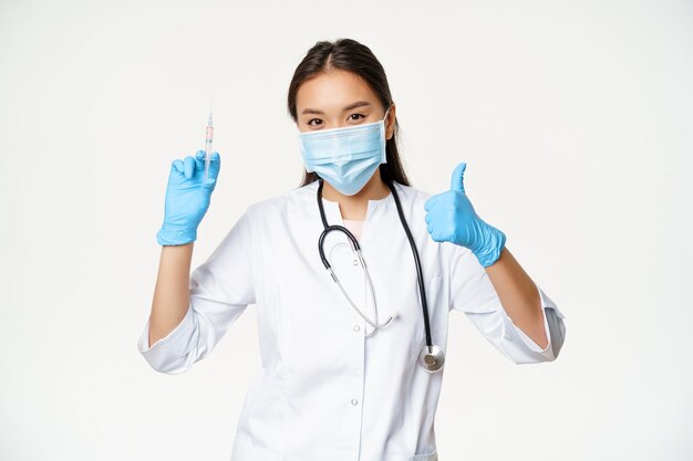 Vaccinazione Covid-19 e assistenza sanitaria. Medico donna asiatica in maschera medica e guanti, tenendo la siringa del vaccino e mostra il pollice in su, sfondo bianco.
