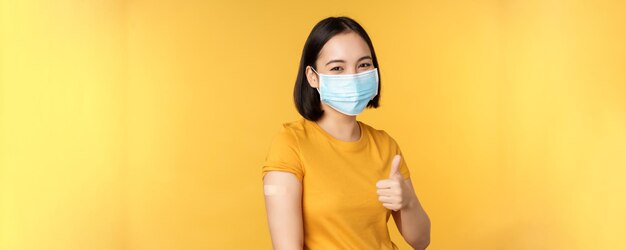 Vaccinazione da covid e concetto di salute Felice ragazza asiatica che mostra i pollici in su indossando maschera medica cerotto sulla spalla ha ottenuto il vaccino contro il coronavirus girato su sfondo giallo