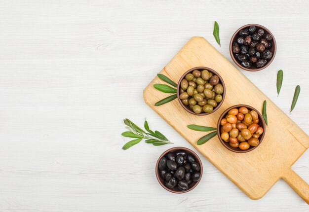 Varietà di olive con foglie di olivo in ciotole di argilla e tagliere su legno bianco, piatto disteso.