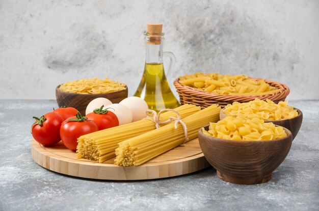 Varietà di pasta cruda con uova, pomodori freschi e bottiglia di olio sulla superficie in marmo.