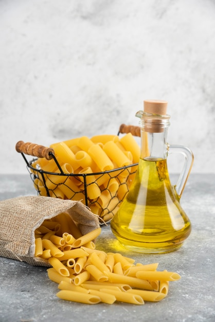 Varietà di pasta in un cesto metallico e una borsa rustica con olio d'oliva sul tavolo grigio.