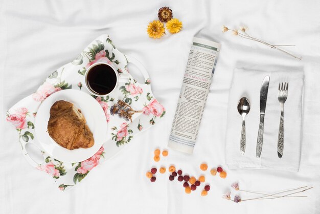 Vassoio colazione floreale; lampone; giornale arrotolato; fiore e posate sul tovagliolo bianco sopra il panno di raso