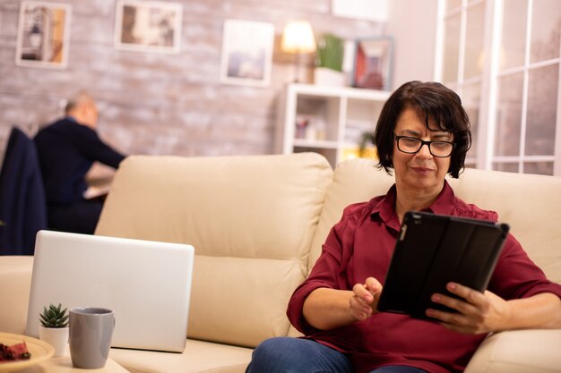 Vecchia donna anziana seduta sul divano e utilizzando un tablet PC digitale in un accogliente soggiorno.