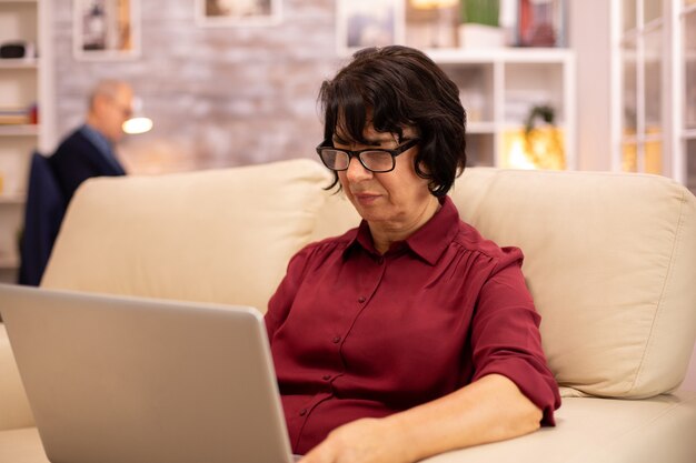 Vecchia donna anziana sul suo divano che lavora su un computer portatile moderno nel suo accogliente soggiorno. Suo marito è in sottofondo