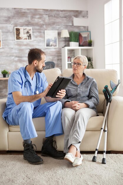 Vecchia donna con le stampelle in casa di cura seduta sul divano ascoltando medico maschio con computer tablet.