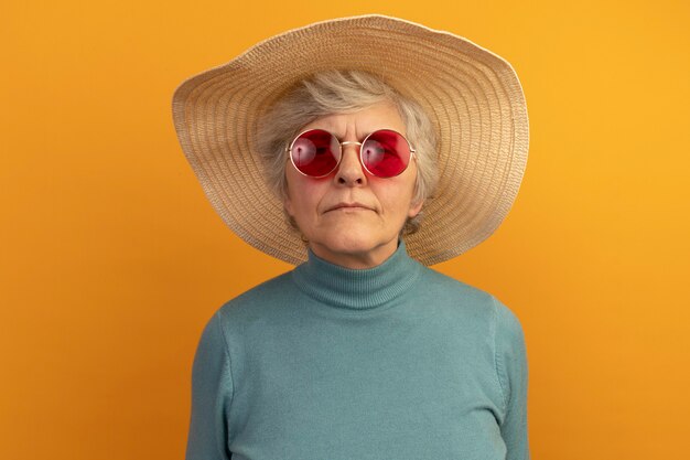 Vecchia donna confusa che indossa un maglione blu a collo alto con cappello da spiaggia e occhiali da sole isolati su una parete arancione