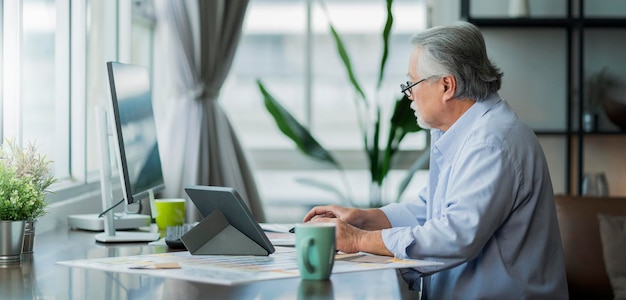 Vecchio asiatico anziano pensionato che lavora a casa mano ise desktop e documento cartaceo alla scrivania in soggiorno interni di casa lavoro maschile senior asiatico a casa