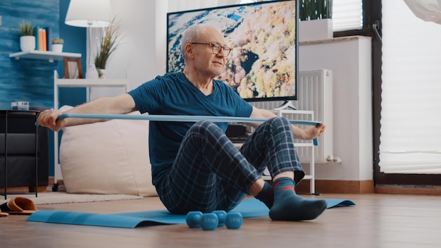 Vecchio che usa la fascia di resistenza per allungare e tirare, seduto su un tappetino da yoga. Addestramento di persona anziana con cintura elastica per esercitare i muscoli delle braccia a casa. Adulto anziano che fa attività fisica