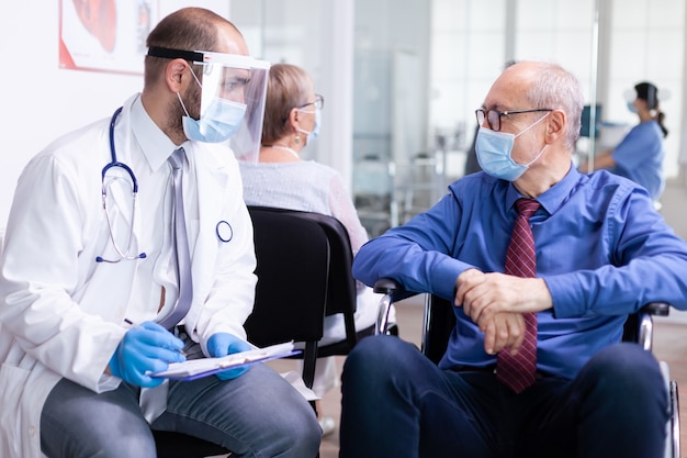 Vecchio invalido con maschera facciale contro l'infezione da coronavirus in sedia a rotelle che discute con il medico nell'area di attesa dell'ospedale