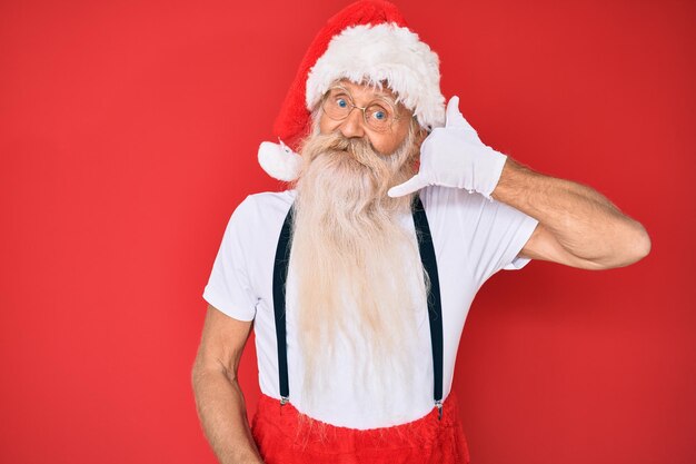 Vecchio uomo anziano con i capelli grigi e la barba lunga che indossa una maglietta bianca e un costume da babbo natale che sorride facendo il gesto del telefono con la mano e le dita come parlare al telefono. comunicare concetti.