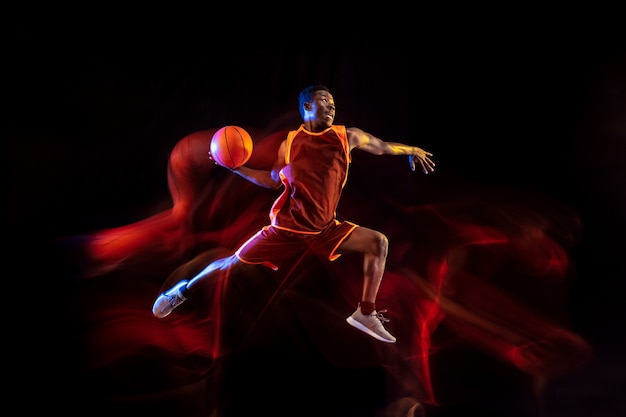 Vedi l'obiettivo. Giovane giocatore di basket afro-americano della squadra rossa in azione e luci al neon su sfondo scuro studio. Concetto di sport, movimento, energia e stile di vita dinamico e sano.