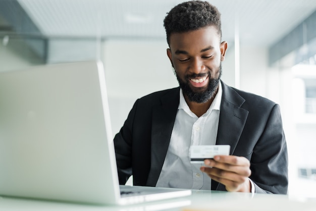 Vendite bancarie su Internet. Riuscito uomo d'affari africano che si siede ad un computer portatile e che tiene la carta di credito a disposizione fino a quando l'uomo d'affari che fa gli ordini tramite Internet