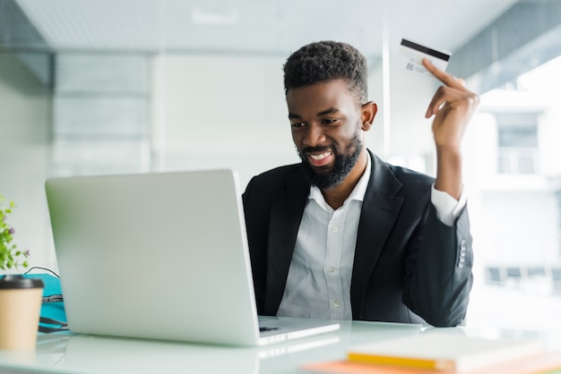 Vendite bancarie su Internet. Riuscito uomo d'affari africano che si siede ad un computer portatile e che tiene la carta di credito a disposizione fino a quando l'uomo d'affari che fa gli ordini tramite Internet