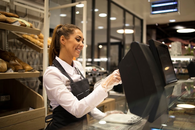 Venditore di panettiere femminile che lavora al computer e che vende pane nel supermercato