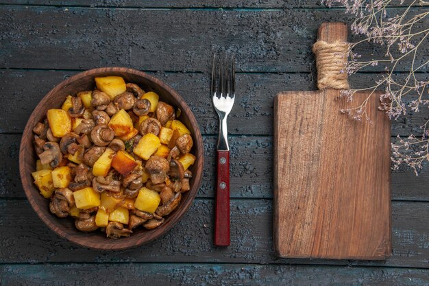 Verdura vista dall'alto con funghi patate e funghi nella ciotola accanto alla forchetta e al tagliere sul tavolo scuro