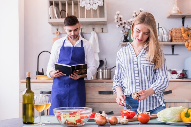 Verdure di taglio della donna mentre uomo che legge il libro di ricette