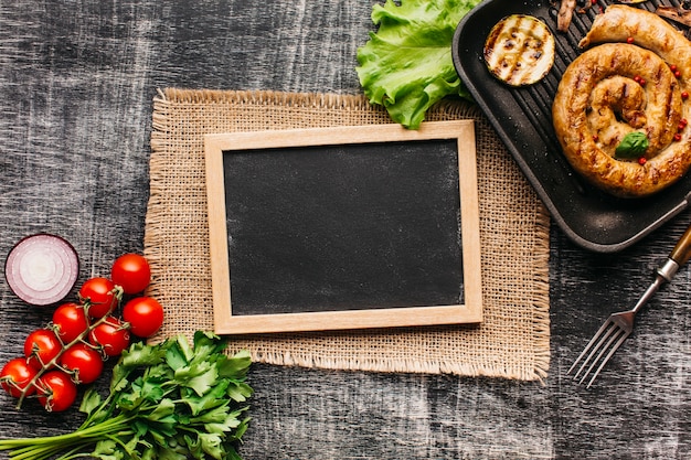 Verdure fresche e spirale salsicce alla griglia con ardesia vuota su sfondo grigio strutturato