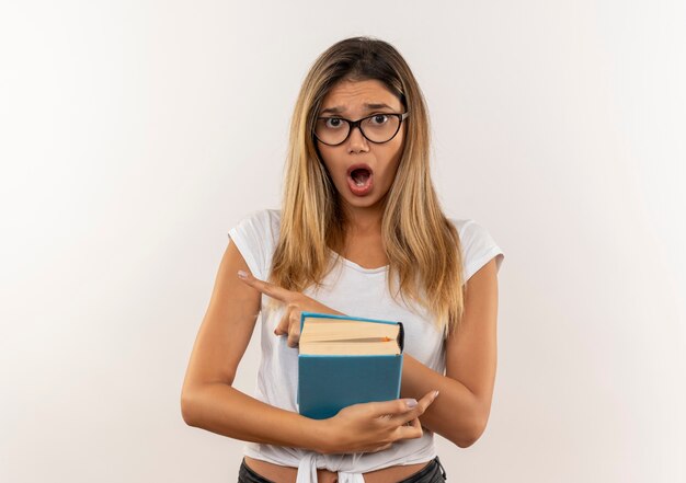 Vetri da portare della giovane ragazza graziosa dell'allievo impressionato e libro della tenuta della borsa posteriore che indica al lato isolato su bianco