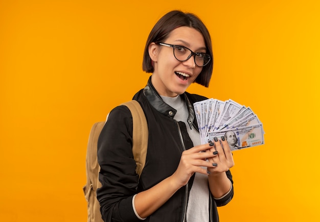 Vetri da portare della ragazza giovane dell'allievo impressionato e denaro della holding della borsa posteriore isolato sull'arancio