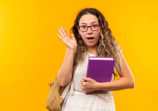 Vetri da portare impressionati della giovane scolara graziosa e libro della tenuta della borsa posteriore che mostra la mano vuota isolata su colore giallo