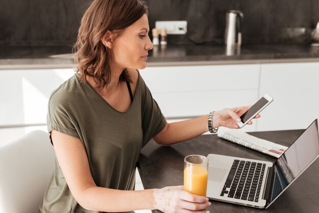 Vew laterale della donna casuale che per mezzo dello smartphone, del computer della compressa e bevendo succo dal tavolo sulla cucina