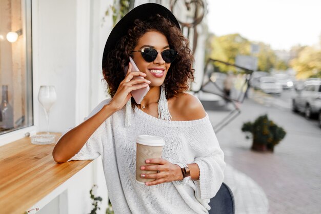 Viaggiare mix gara donna in elegante abito casual rilassante all'aperto nel caffè della città, bere caffè e chattare con il telefono cellulare. Indossa accessori e occhiali da sole alla moda.