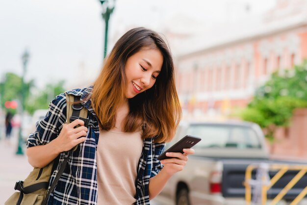 Viaggiatore con zaino e sacco a pelo turistico della donna asiatica che sorride e che usando smartphone che viaggia da solo