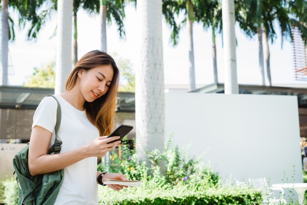 Viaggiatore con zaino e sacco a pelo turistico della donna asiatica che sorride e che usando smartphone che viaggia da solo