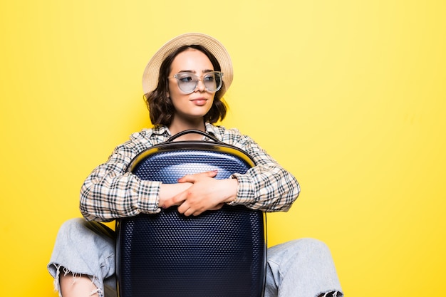 Viaggiatore femminile elegante felice in cappello di paglia in piedi con borsa con ruote