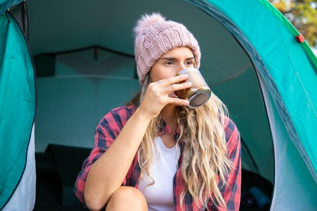 Viaggiatore femminile seduto in tenda e bere il tè. Cappello da portare della donna bionda caucasica, campeggio sulla natura, distogliere lo sguardo e godersi la vista. Concetto di turismo, avventura e vacanze estive con lo zaino in spalla